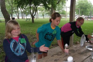 Part of the Food Science workshop at the Adventure Day Camp found these 4-H members experiencing inertia and gravity. Madison Mandel, Reese Hansen and Sami Linder watching the eggs experience gravity and drop safely in the glass of water after they provided the unbalanced force to change inertia (hitting) the pie tin and toilet paper tube out from underneath them. Photo Credit/Mary Loftis, Extension Assistant.