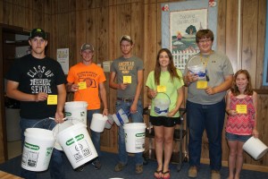 Keeping livestock properly watered takes a lot of water and effort if you are carrying it to your animals as these Burt County 4-H members learned. They are pictured with at least part of the buckets they would have to carry to the animal they were assigned as part of the Livestock Quality Assurance Training last Sunday evening in Oakland. Pictured are: Bailey Fleischman of Tekamah showing part of the 23 gallons of water a 1150 lb. steer would need at 90 degrees; Bryce Uhing of West Point with enough water for a 700 lb steer at 50 degrees; Thomas Hennig of Tekamah with the 6 gallons of water he’d need for a 200 pound hog at 100 degrees; Megan Olson of Tekamah with the much smaller amount she’d need for a 40 lb. lamb at 50 degrees; Blaine Olson of Tekamah with water for his 120 lb. lamb at 80 degrees and Sydney Olsen of Lyons with enough water for a 120 lb. pig at 50 degrees. The rule of thumb in calculating water use is one gallon per 100 lbs. of animal plus more for heat and stress situations. Photo Credit/Mary Loftis